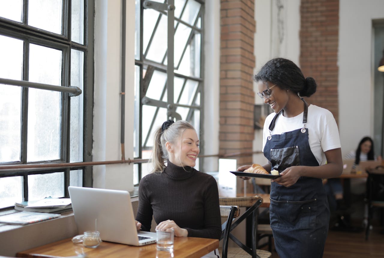 Friendly interaction in a café with a waitress serving a customer using a laptop. Bright and welcoming atmosphere.